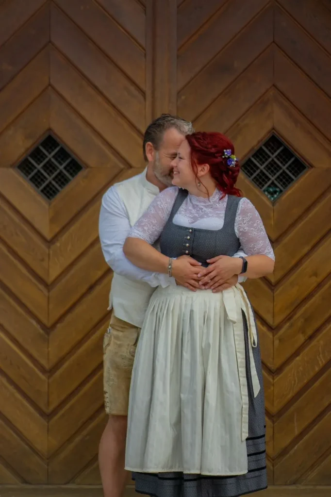 Brautpaar in Trachtenmode gekleidet. Brautpaar steht vor einem Holztor. Bräutigam steht dabei hinter der Braut und umarmt diese. Fotografiert von hochzeitsfotograf-bamberg.net