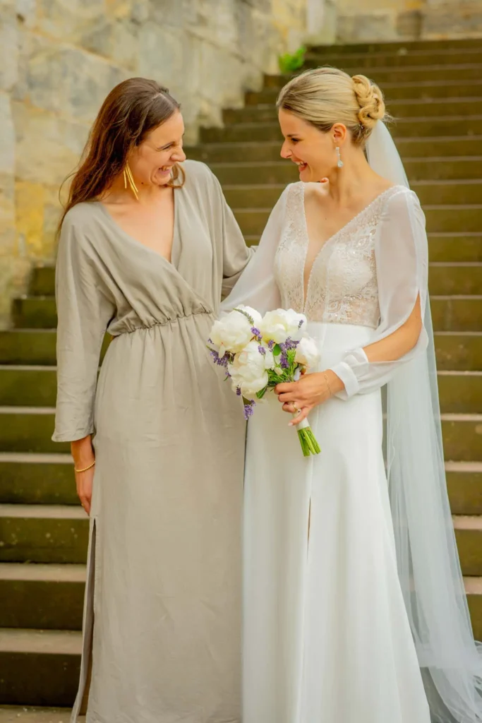 Braut in weißem Hochzeitskleid und langem Schleier steht neben ihrer Trauzeugin. Beide schauen sich lächelnd an. Fotografiert von hochzeitsfotograf-bamberg.net