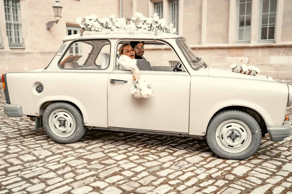 Brautpaar fährt im Trabi. Braut hält den Blumenstrauß aus dem Fenster und lächelt in die Kamera. Fotografiert von hochzeitsfotograf-bamberg.net