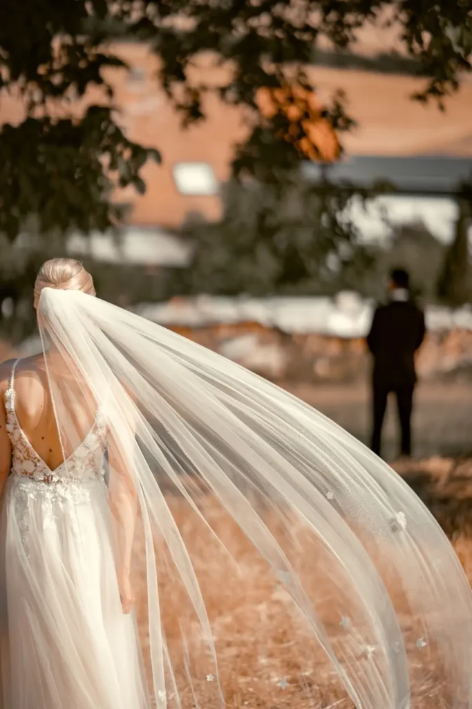 Braut steht mit dem Rücken zum Fotografen und sieht in Richtung des Bräutigams. Der weiße Schleier wird vom Wind getragen. Fotografiert von hochzeitsfotograf-bamberg.net