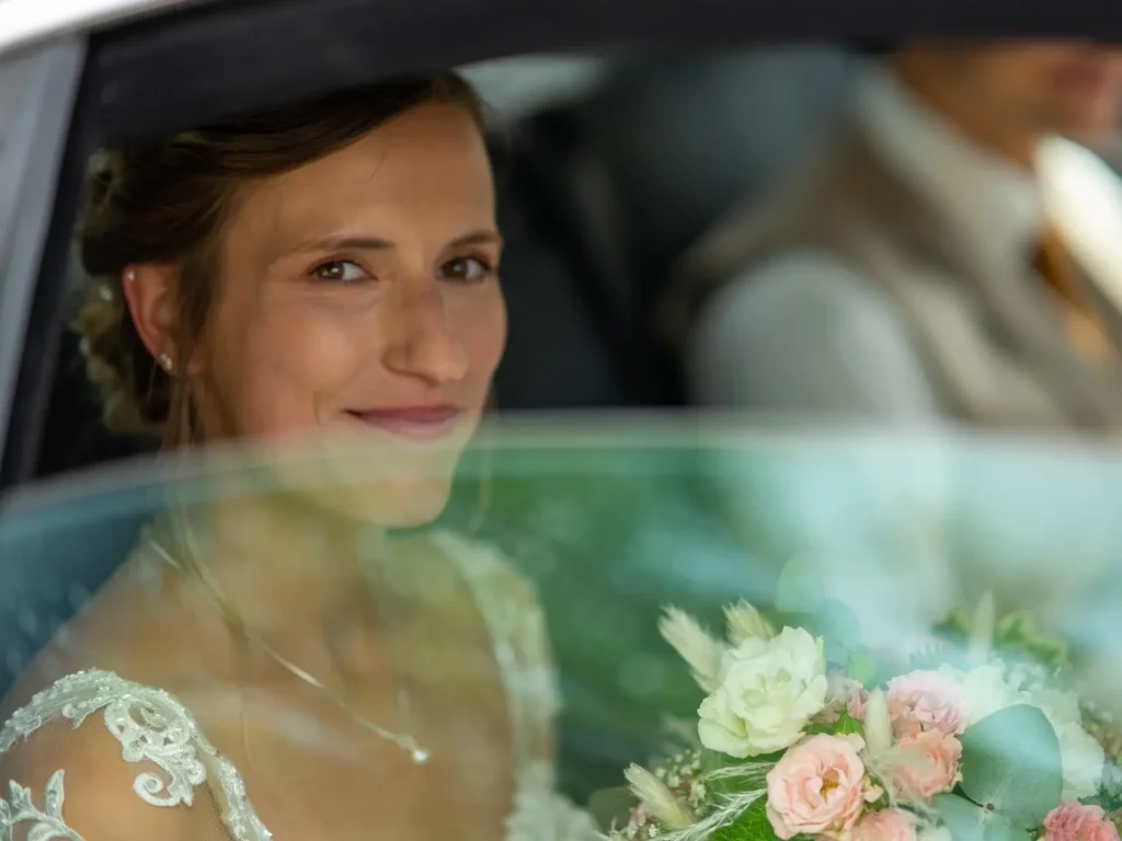 Braut sitzt im Hochzeitsauto und lächelt aus dem halb geöffneten Fenster in die Kamera. Fotografiert von hochzeitsfotograf-bamberg.net
