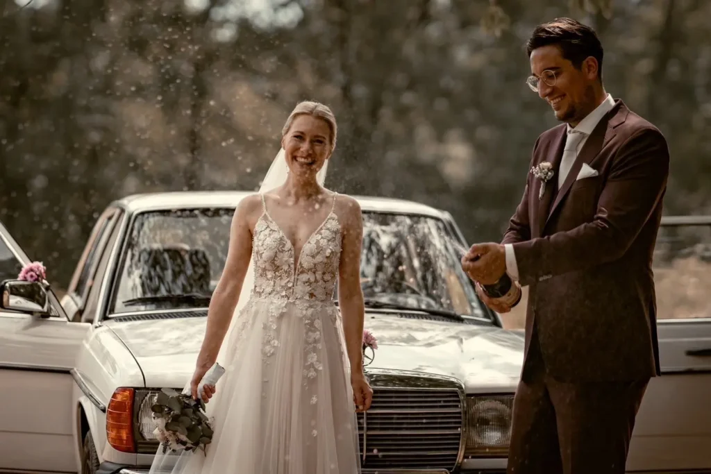 Brautpaarshooting in Bamberg. Brautpaar steht vor dem Hochzeitsauto. Bräutigam spritzt mit einer Sektflasche. Fotografiert von hochzeitsfotograf-bamberg.net.