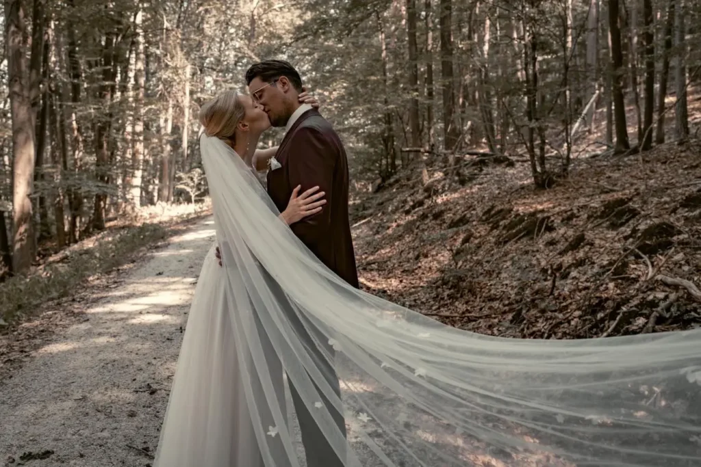Brautpaar steht auf einem Waldweg und küsst sich. Der lange weiße Schleier wird vom Wind getragen. Fotografiert von hochzeitsfotograf-bamberg.net