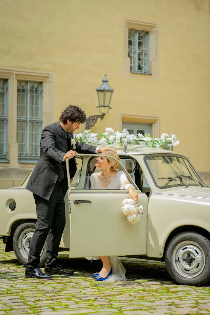 Brautpaarshooting. Bräutigam steht auf der Beifahrerseite des Trabis, welcher als Hochzeitsauto fungiert. Die Beifahrertür steht offen. Die Braut sitzt auf dem Beifahrersitz und lächelt dem Bräutigam zu. Fotografiert von hochzeitsfotograf-bamberg.net