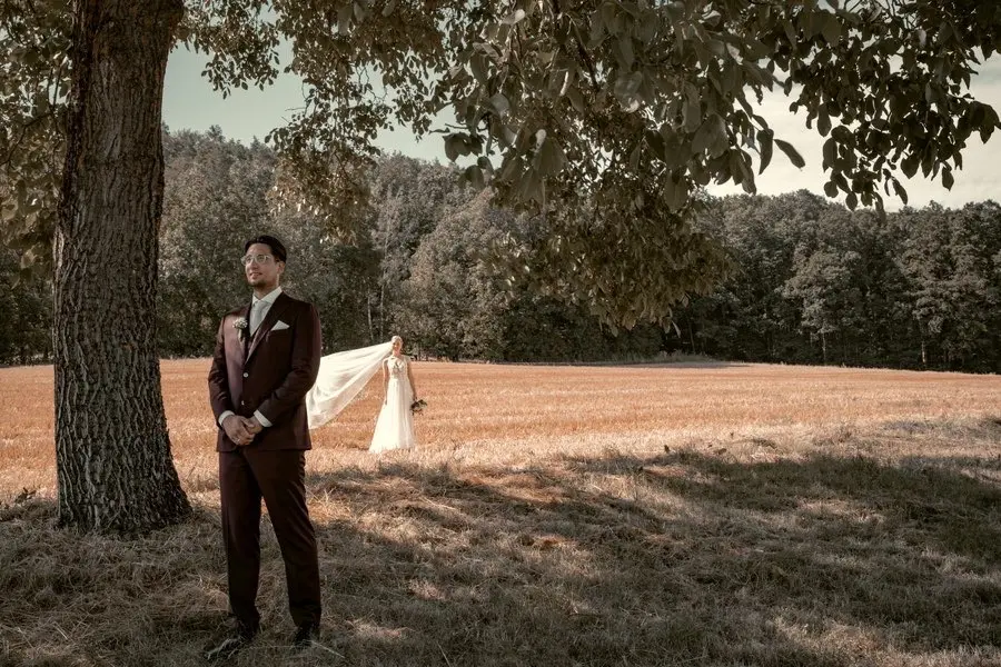 Hochzeitsfotografie. Bräutigam steht unter einem Baum. Die Braut steht hinter ihm auf einem Feld. Fotografiert von hochzeitsfotograf-bamberg.net