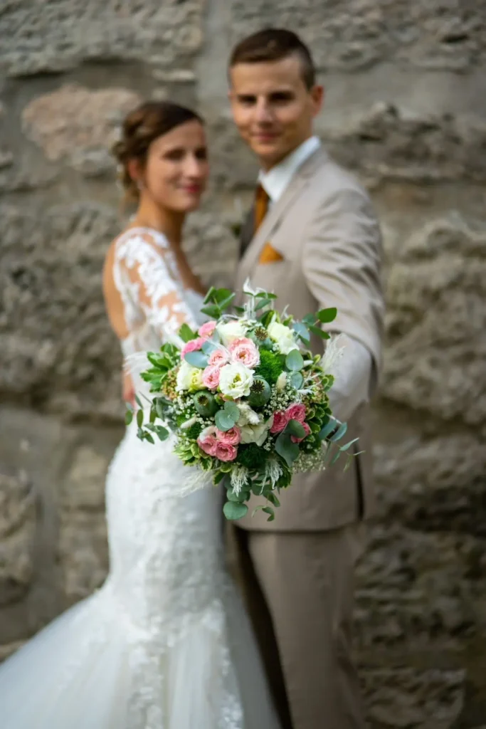 Brautpaar steht Brust an Brust und hält den Blumenstrauß in die Kamera. Blumenstrauß ist scharf zu sehen. Das Brautpaar ist unscharf. Fotografiert von hochzeitsfotograf-bamberg.net