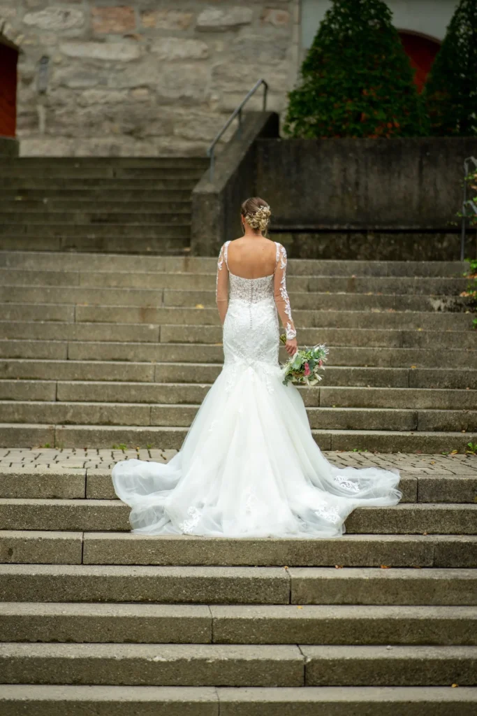 Braut in weißem Hochzeitskleid. Steht mit dem Rücken zum Fotografen. Den Brautstrauß hält sie in der rechten Hand. Fotografiert von hochzeitsfotograf-bamberg.net