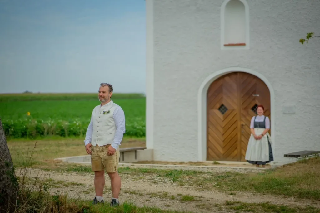 First Look Shooting. Braut und Bräutigam stehen vor einer kleine Kapelle. Bräutigam wartet darauf, dass ihn die Braut an der Schulter antippt. Fotografiert von hochzeitsfotograf-bamberg.net