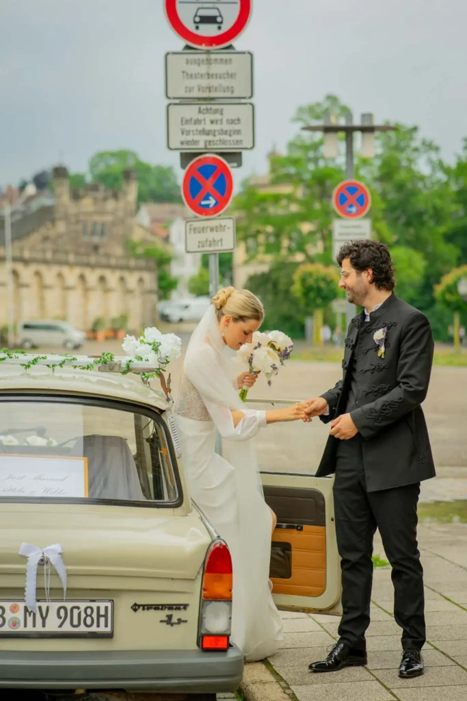 Bräutigam hilft der Braut aus dem Trabi auszusteigen. Braut trägt ein weißes Brautkleid mit langem Schleier. Brutigam trägt einen schwarzen Anzug. Fotografiert von hochzeitsfotograf-bamberg.net